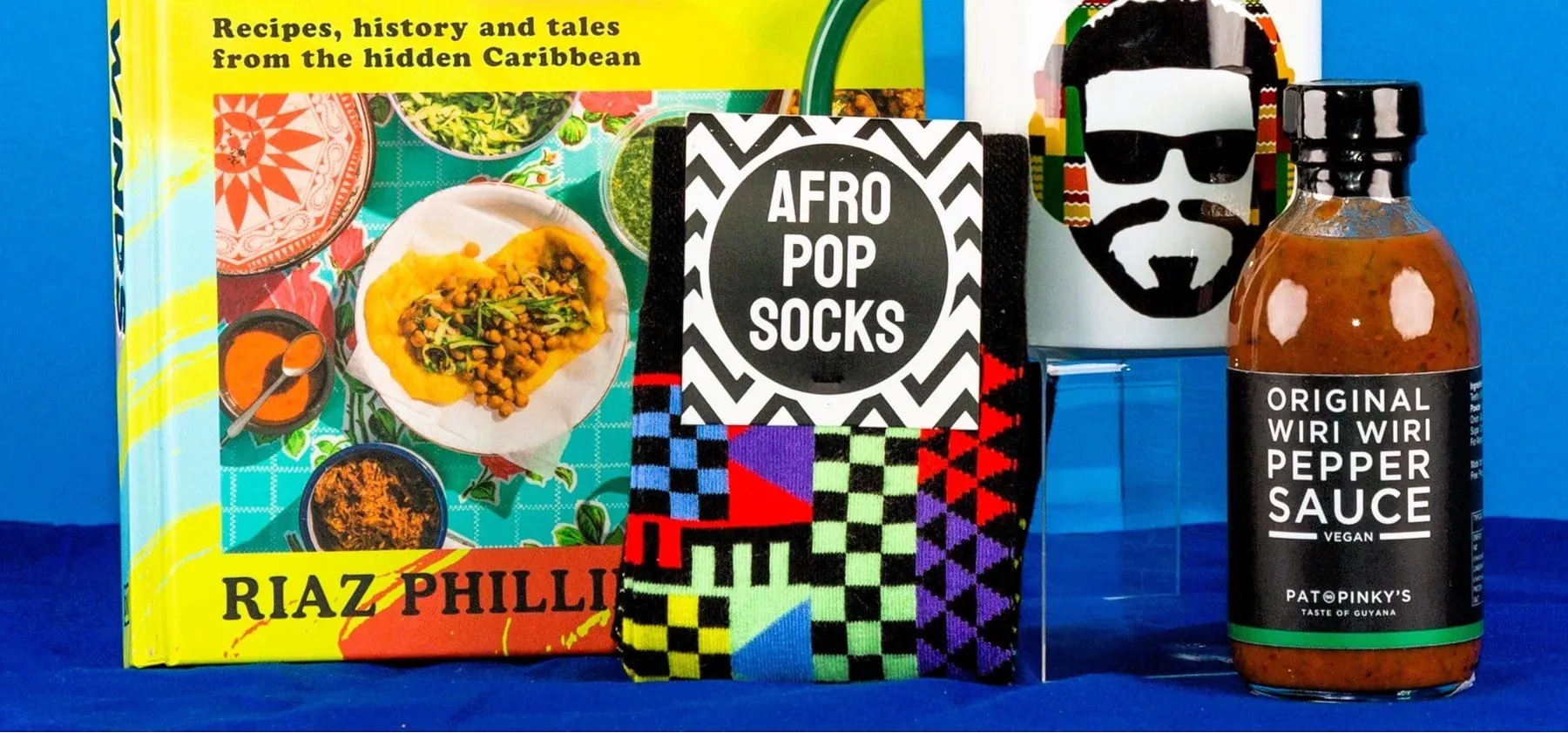 Book titled 'East Winds' by Riaz Phillips, a bag labeled 'Afro Pop Socks', and a bottle of 'Original Hot Wing Pepper Sauce' on a blue background.