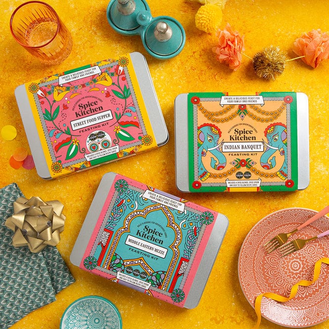 Three colorful Spice Kitchen packaging on a yellow surface with decorative items.